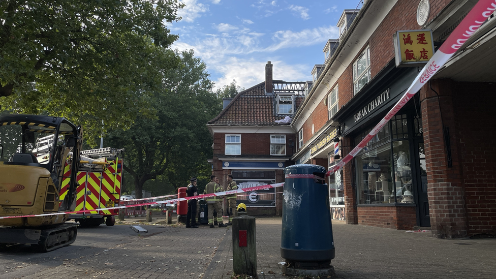 Fire and rescue tape across a parade of shops, with a fire engine in background and burnt out roof of a flat above a shop