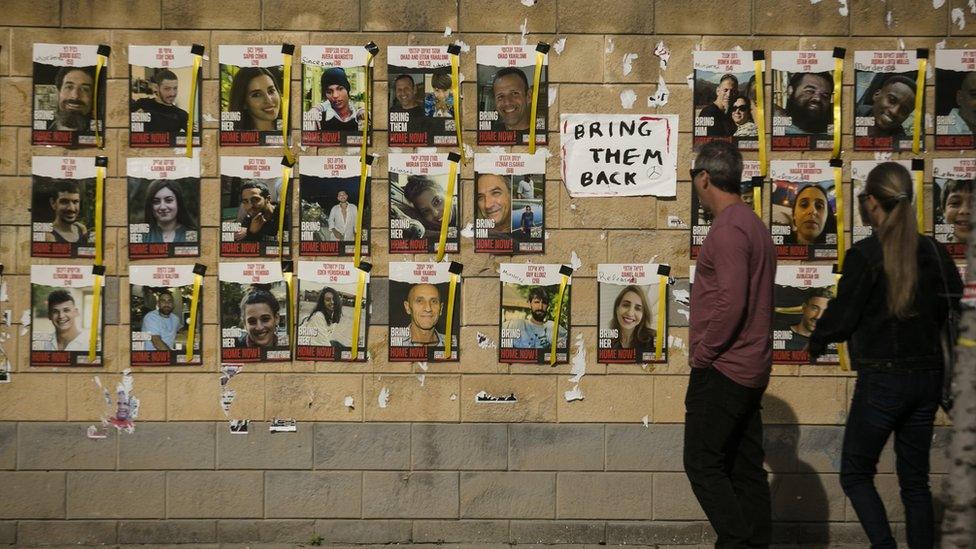 People walk past posters of Israeli hostages held in Gaza and a sign saying "Bring them back", on a wall in Tel Aviv, Israel (20 December 2023)