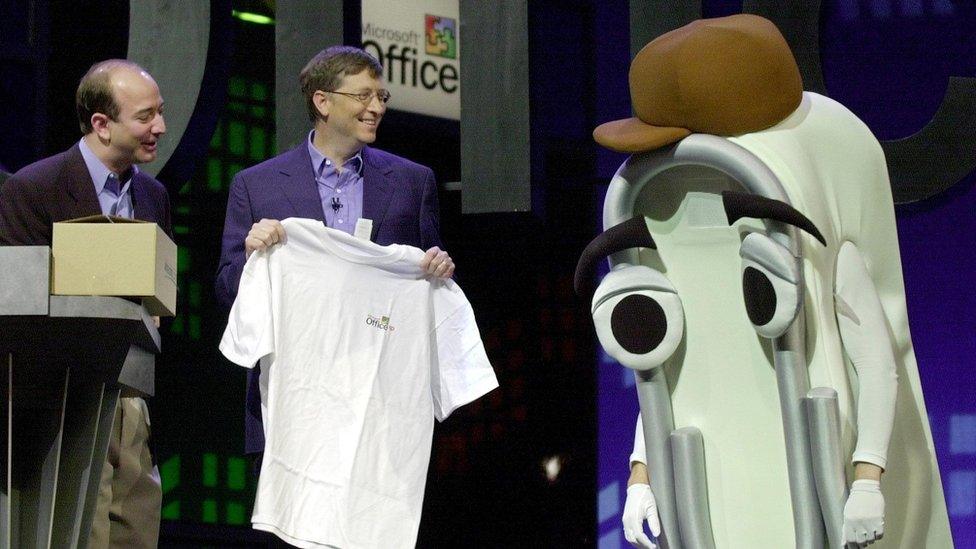 Bill Gates smiles holding a white t-shirt next to an actor wearing a giant Clippy foam suit