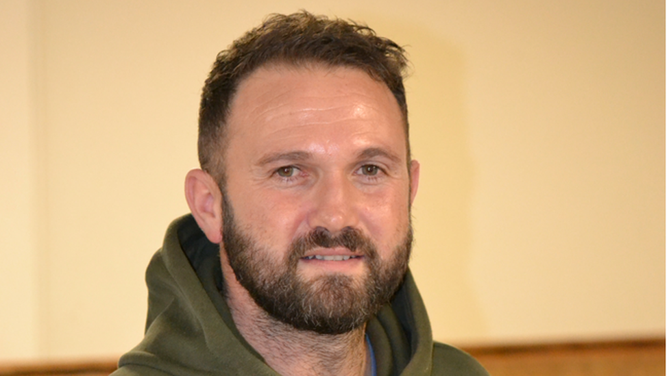 Dark haired man with beard, wearing a green hooded top