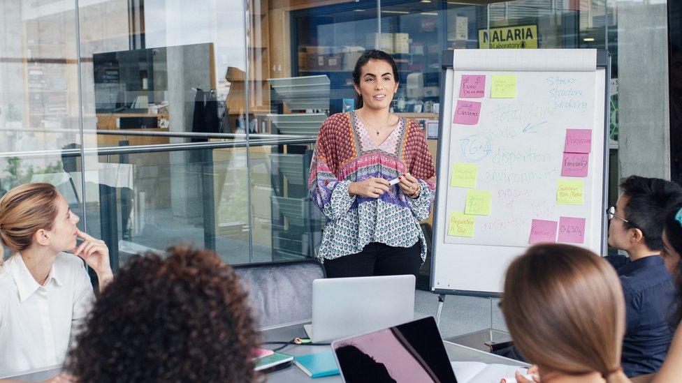 A Spanish female is heading up a business meeting to female colleagues in a glass cubicle office