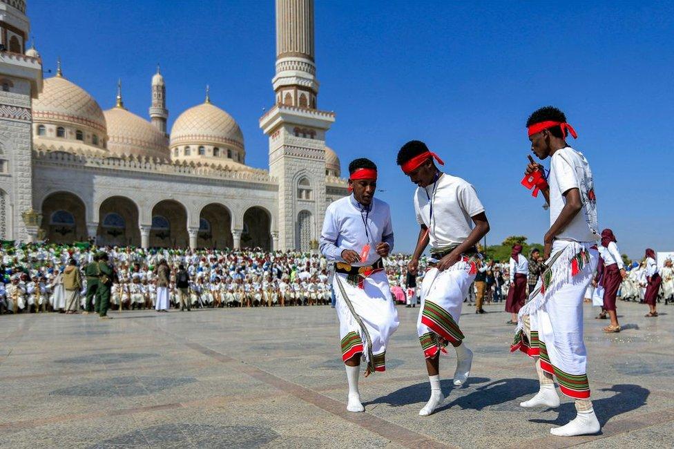 Three men do a footwork dance. In the background there are scores of people, and a large church.