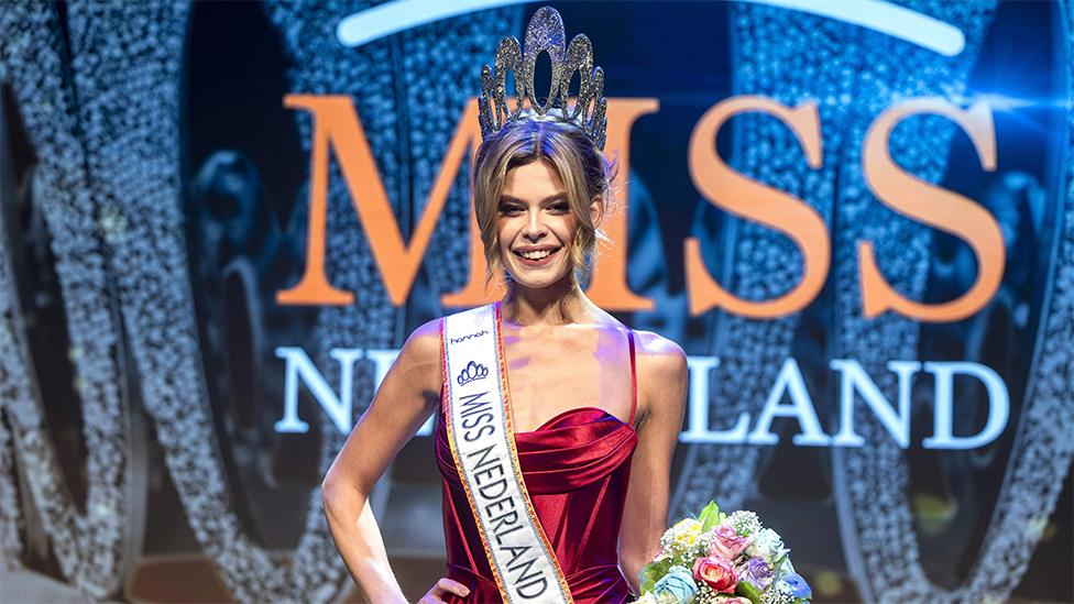 Rikkie Valerie Kolle after being crowned Miss Netherlands on stage. She is wearing a red dress and has a white sash around here around here saying Miss Netherlands 2023. She has a big silver tiara on her head and flowers in her left hand. The background has a sign in orange and white saying Miss Netherland