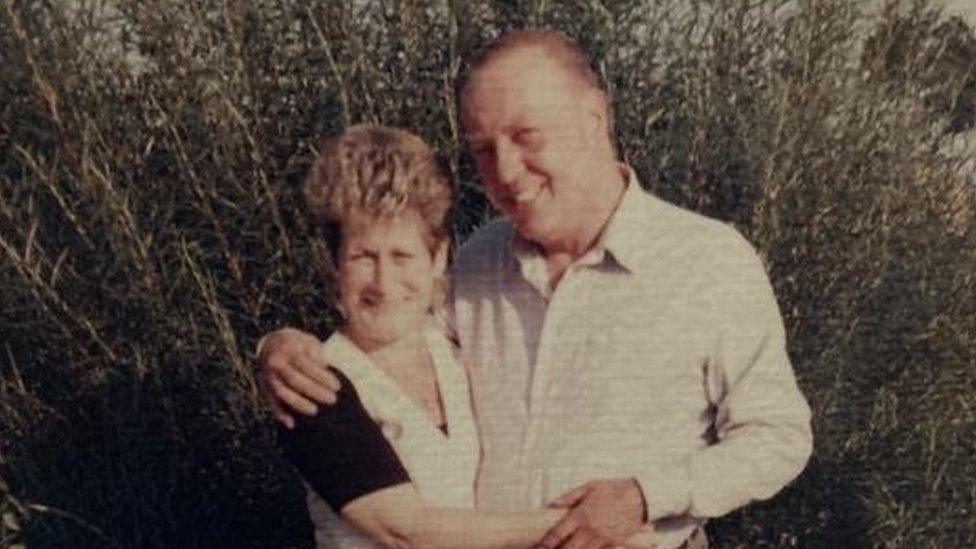 A photograph of Ron with his wife Trish holding each other. They are outside and standing in front of a shrub.