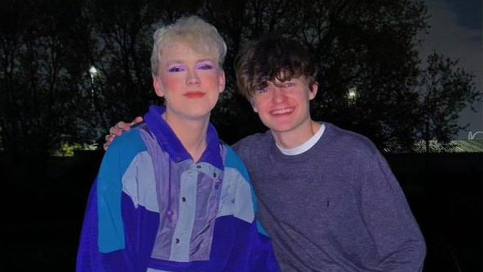 Aaron and Archie smiling at the camera. Aaron is wearing a blue and purple shirt, and purple eye make-up. Archie is wearing a grey jumper.