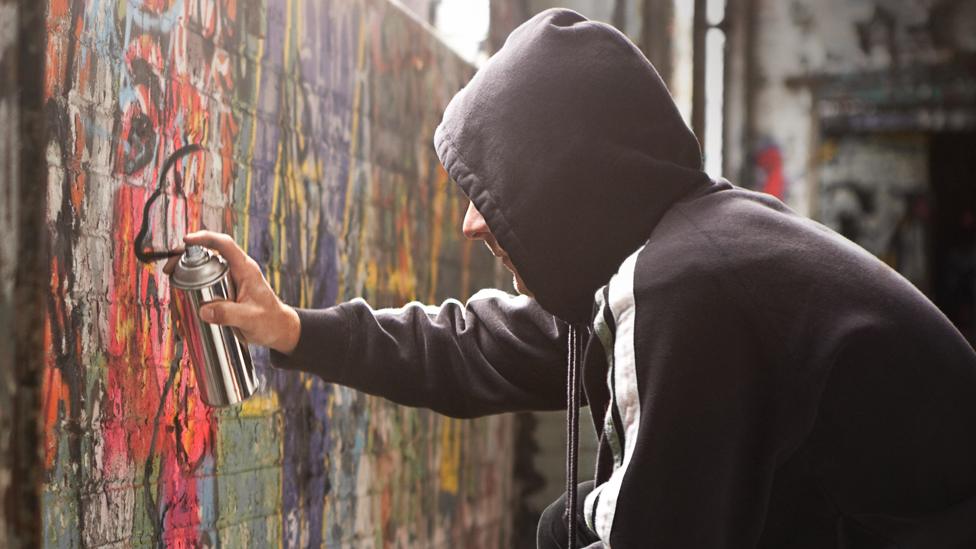 Stock image of a man with a hood spraying black paint on a wall of graffiti