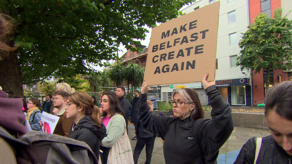 A woman holds up sign that reads: MAKE BELFAST CREATE AGAIN