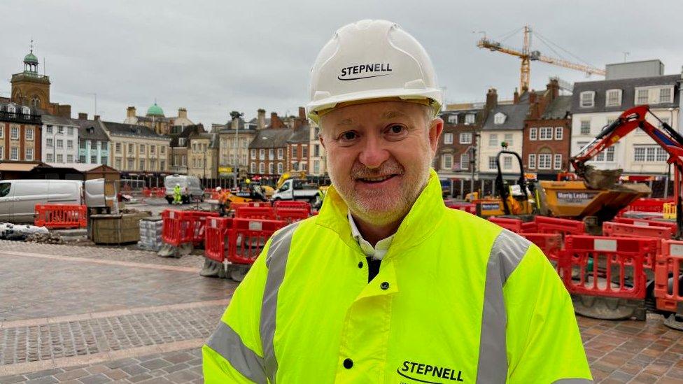 Bearded man in yellow hi-viz and hart hat stands in front of fenced off part of market square