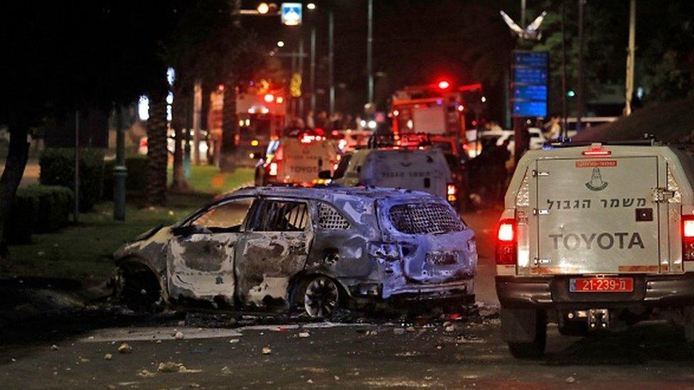 Israeli border police vehicles drive past an extinguished burnt vehicle in Lod near Tel Aviv