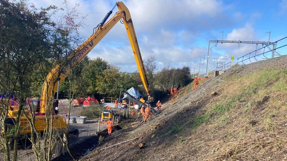 Large yellow digger at work on an embankment. Workers in orange hi-viz are visible