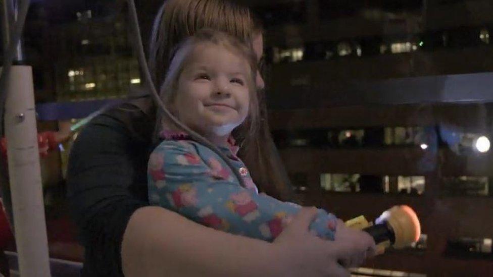 A young girl smiles as she sits on her mum's lap in front of a window from where twinkling lights can be seen down below