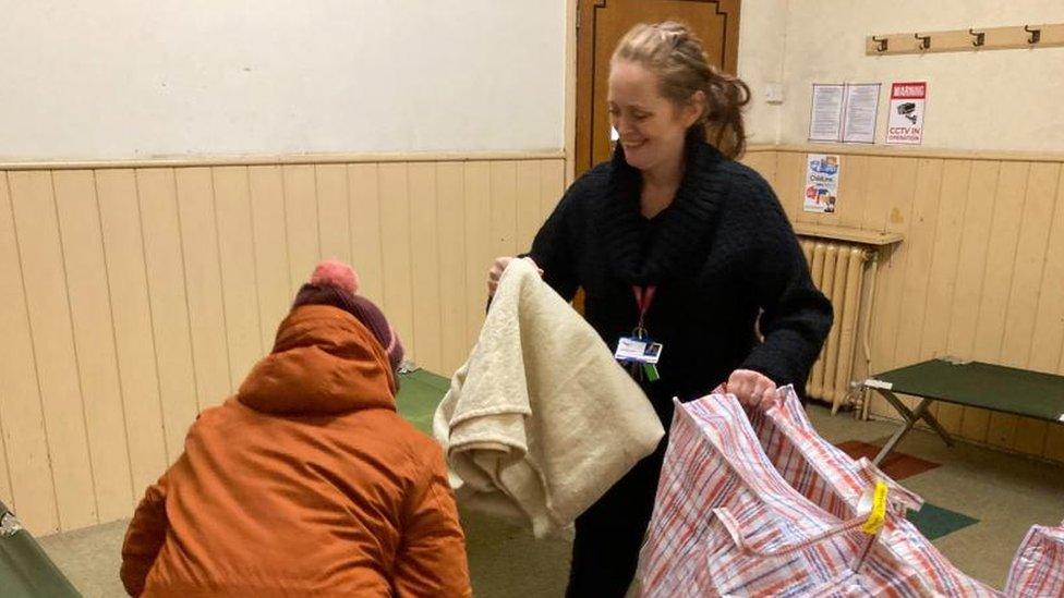 Woman wearing lanyard takes bed sheets from a large bag while a woman in a orange raincoat sits on a bed.