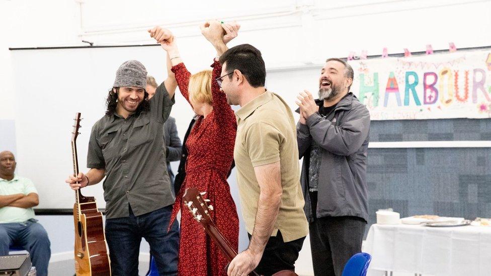 Claire Garrett CEO of The Harbour Project holding up the arms of Mauricio from El Salvador and Amirreza from Iran as they are applauded for their acoustic performance