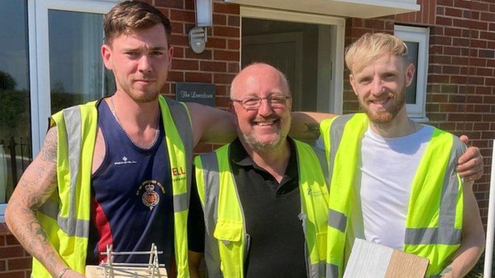 Military veterans Jack Aldis, Kevin Chapman and Jamie Powell stand with their arms around one another holding their construction style trophies presented to celebrate the end of their training