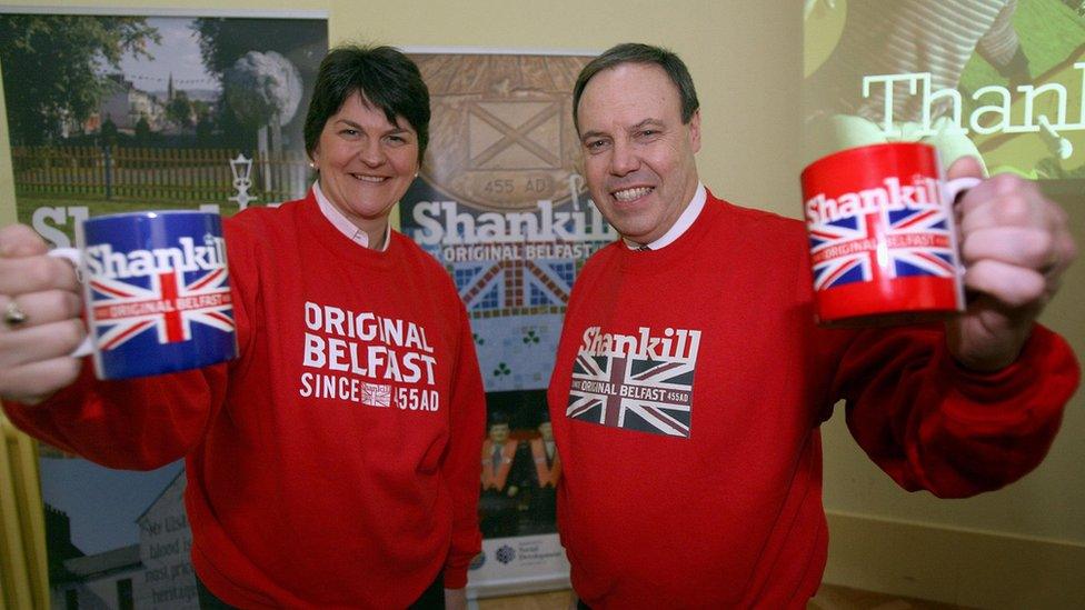 Arlene Foster and Nigel Dodds wearing Shankill-branded sweaters and holding Shankill-branded mugs in 2008