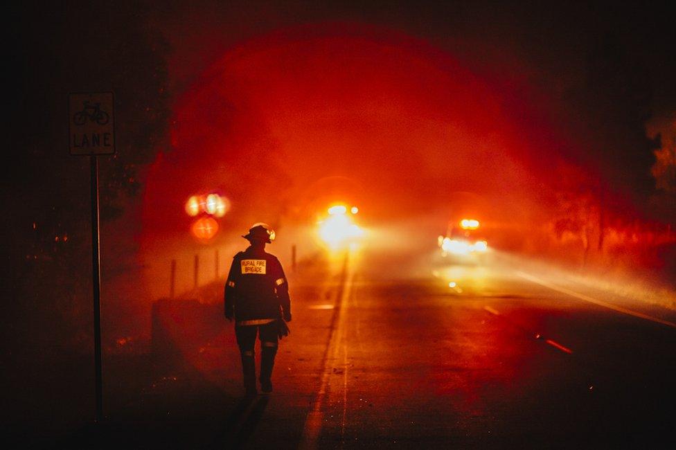 A firefighter walks along a roadside, framed by the red flow of distant lights