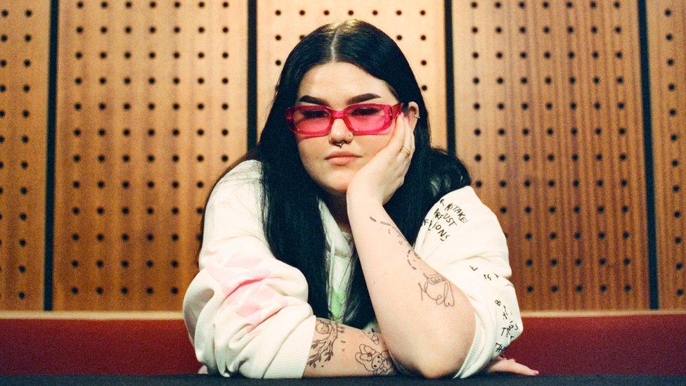 Woman with long dark hair and wearing white top with red glasses sits with her chin in her palm