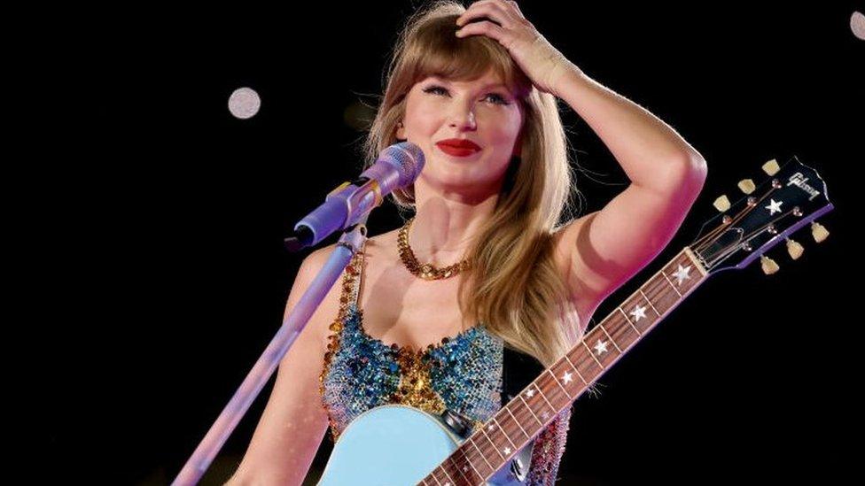 Taylor Swift on stage, she is wearing a blue leotard and has a blue guitar. Her hand is in her straight, blonde hair as she smiles