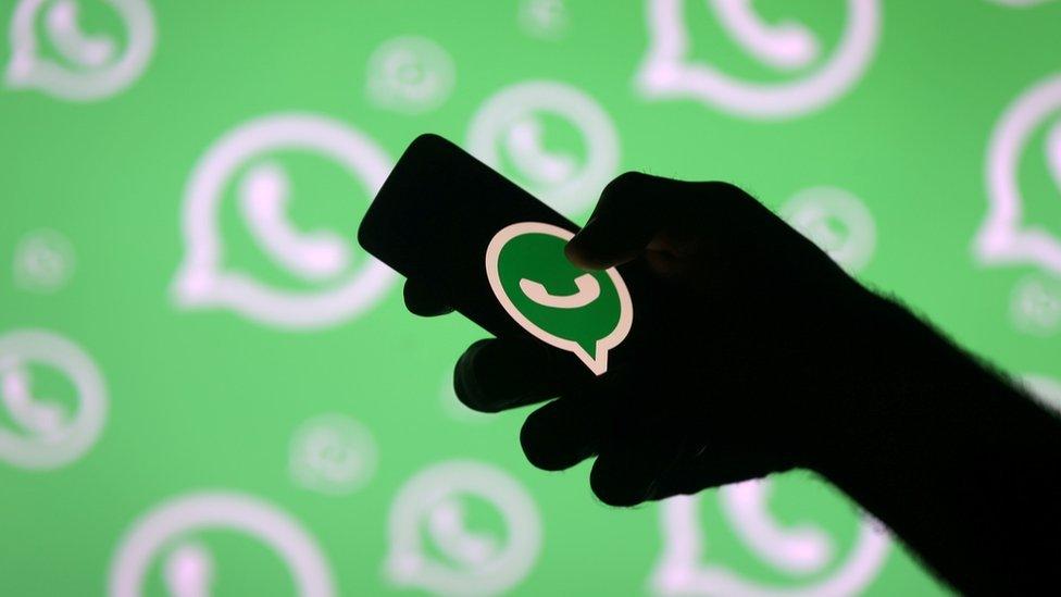 A silhouetted hand useds a smartphone bearing the WhatsApp logo