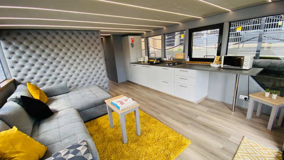 A kitchen lounge interior room inside the floating showhome with sink, counter, microwave, sofa, and luxurious looking quilted wall with wooden flooring