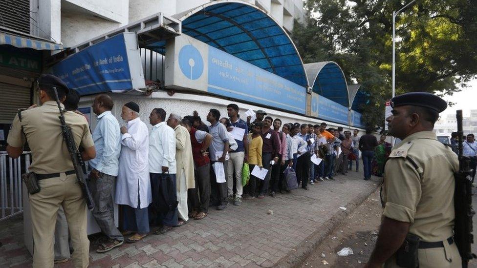 Indian policemen stand guard as people queue up outside a bank to exchange and deposit Indian currency of rupees 500 and 1000 denominations in Ahmadabad, India, Thursday, Nov. 10, 2016.