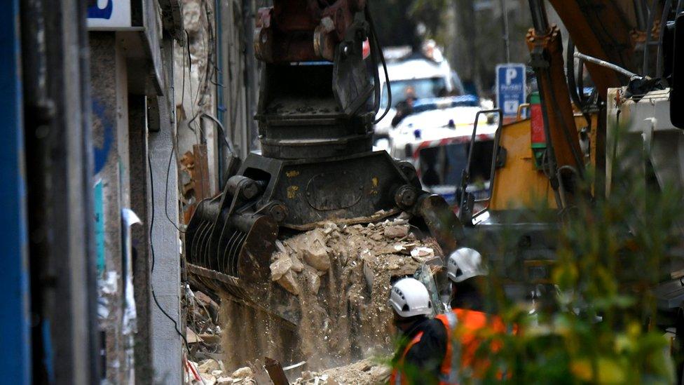 A large digger picks up large amounts of debris spilling dust