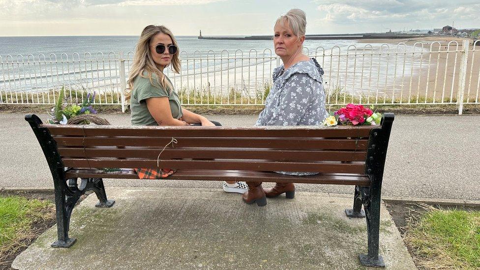 Two women sit on a bench overlooking the sea