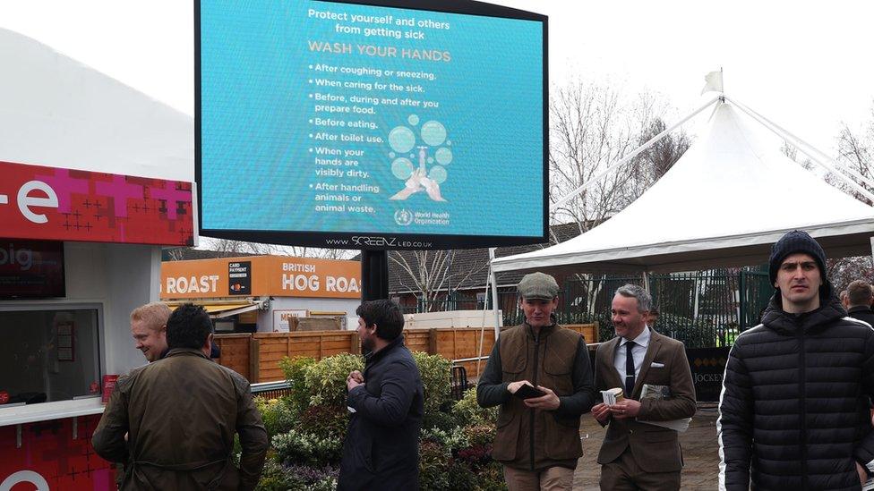 Racegoers pass a sign referencing how to protect yourself and others from getting sick following the Coronavirus outbreak ahead on day one of the Cheltenham Festival at Cheltenham Racecourse