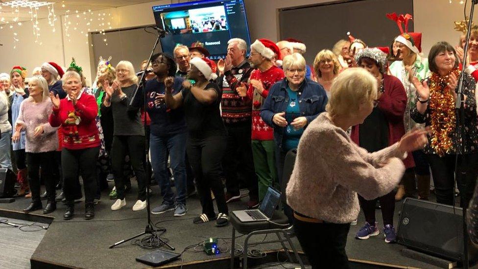 A group of older people. many wearing Christmas hats, sing and dance