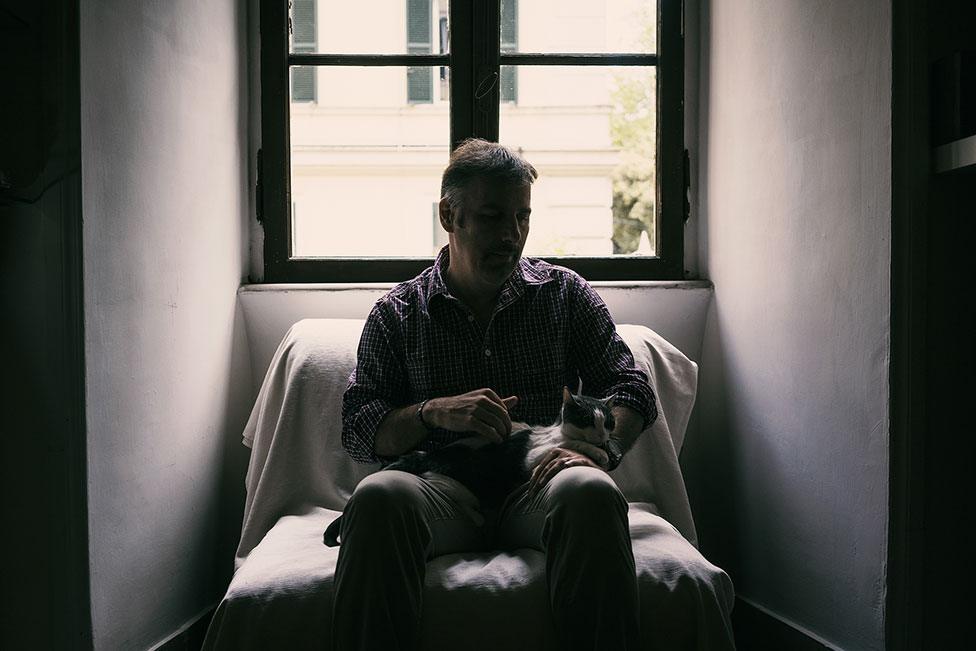 A man sits in front of a window whilst holding a cat in his lap