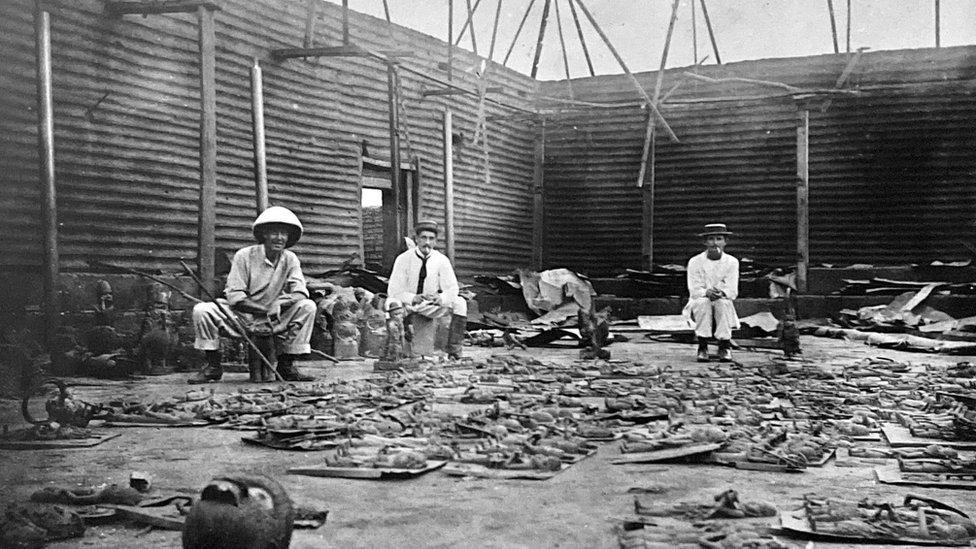 Interior of the Royal Palace during looting in February 1897, showing Captain Charles Herbert Philip Carter, EP Hill, and an unnamed man, with bronzes laid out on the floor.