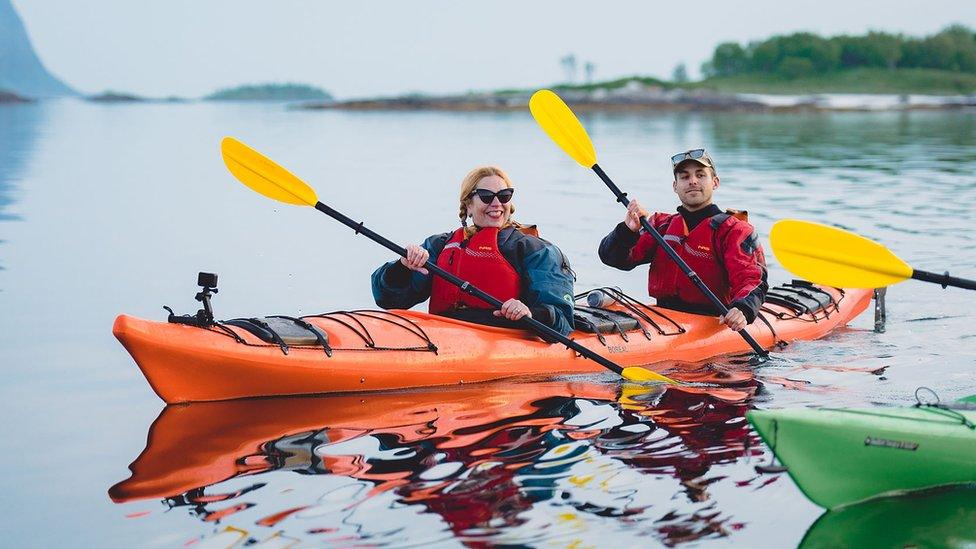 Kazzy in front paddling a double kayak on a bay with mountains in the distance