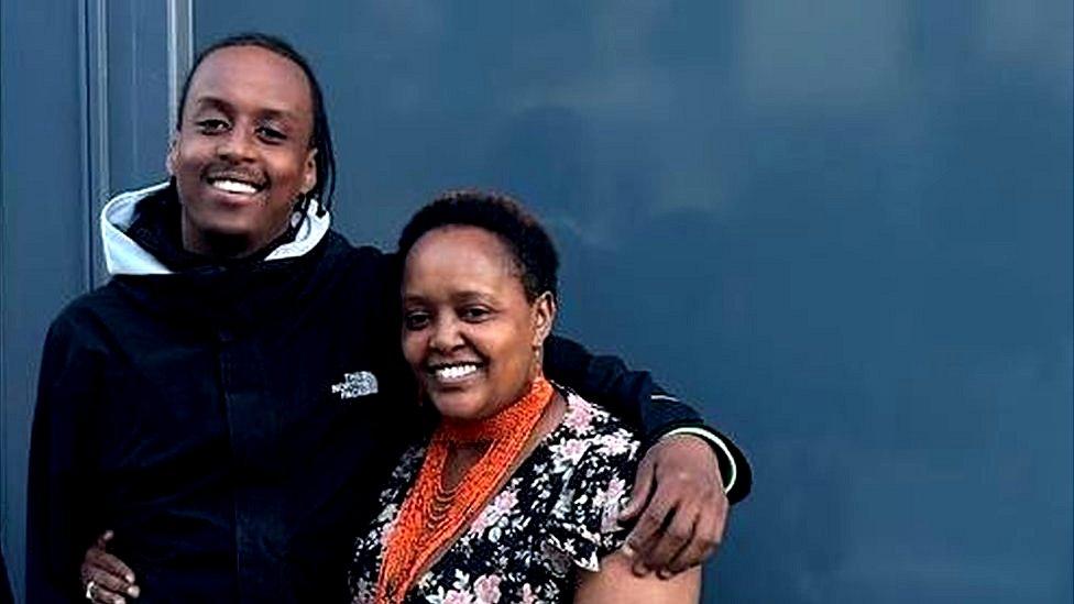 A young black man and a middle aged black woman with their arms around each other smiling