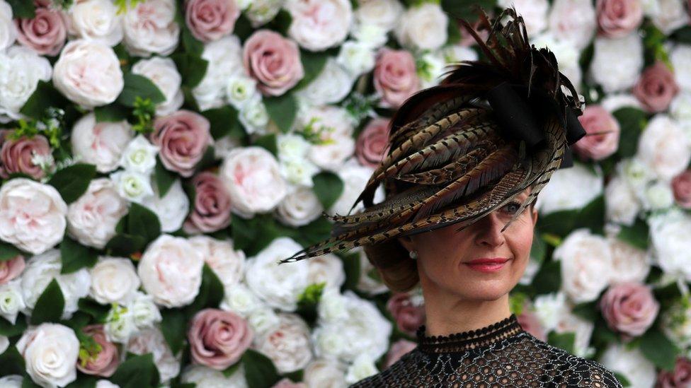 A woman wearing a hat covered in bird feathers stands in front of a wall of roses