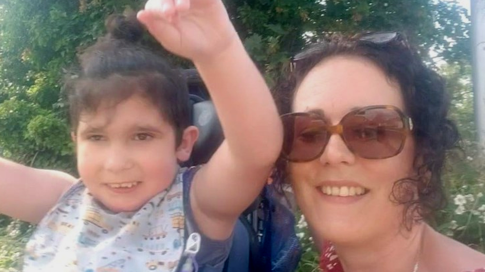 6 year old boy with dark brown hair waving his arms, next to mum with curly brown hair wearing sunglasses