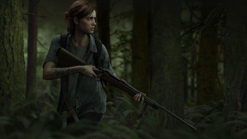 Ellie stands with a gun, looking at something off camera in a green forest