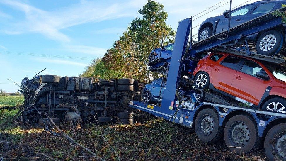 The front part of the car transporter turned over in a field, with the back still attached filled with new cars and just about standing still.