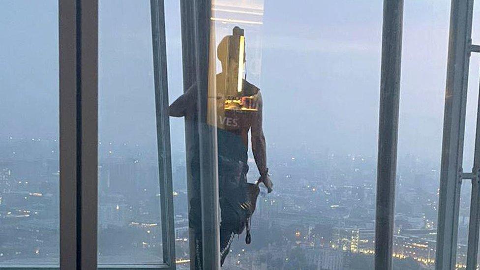 A man looks into the window of the Shard with the skyline of London City behind him