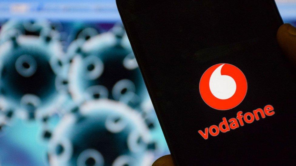 A mobile phone is seen with the Vodafone logo on it in front of an illustration of the coronavirus