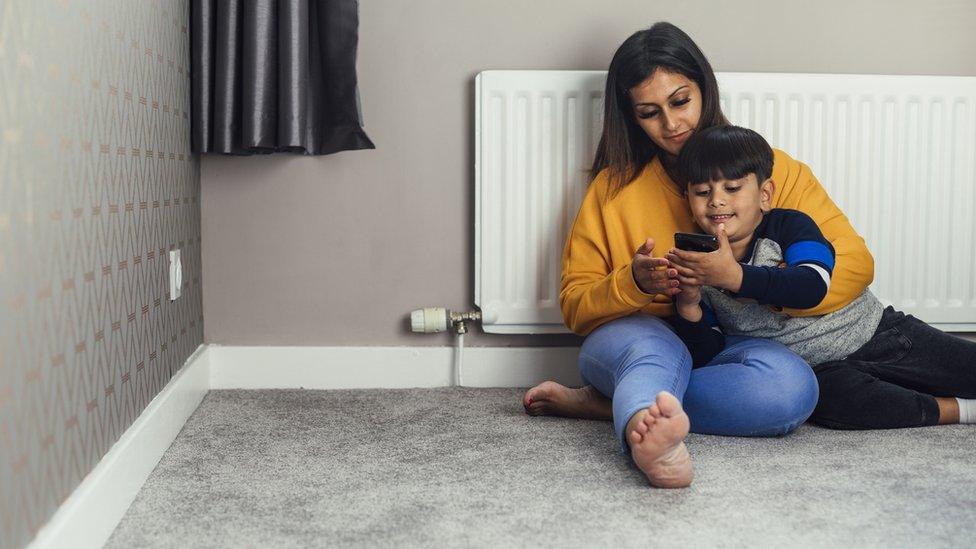 a woman and a child sitting together with their backs to a radiator looking at something on a mobile phone