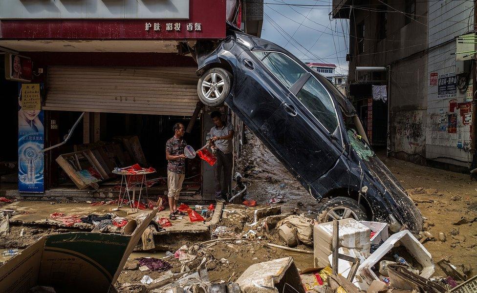 People stand below a car angled 45 degrees, with its rear wheels several metres in the air, in a damaged shopping street filled with debris, on 12 July 2016 in Fuijan, China.