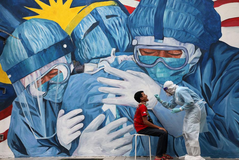 A doctor collects a swab sample from a man, in front of a large mural depicting people wearing personal protective equipment