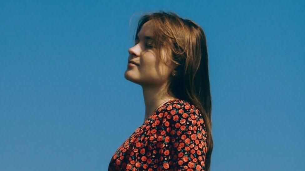 Anna Prasek, known as anMari. Anna is a 22-year-old white woman and stands sideways, her long brown hair worn loose down her back. She has her eyes closed and her head tilted up. She is pictured outside against a blue sky wearing a black dress covered in a red floral pattern.