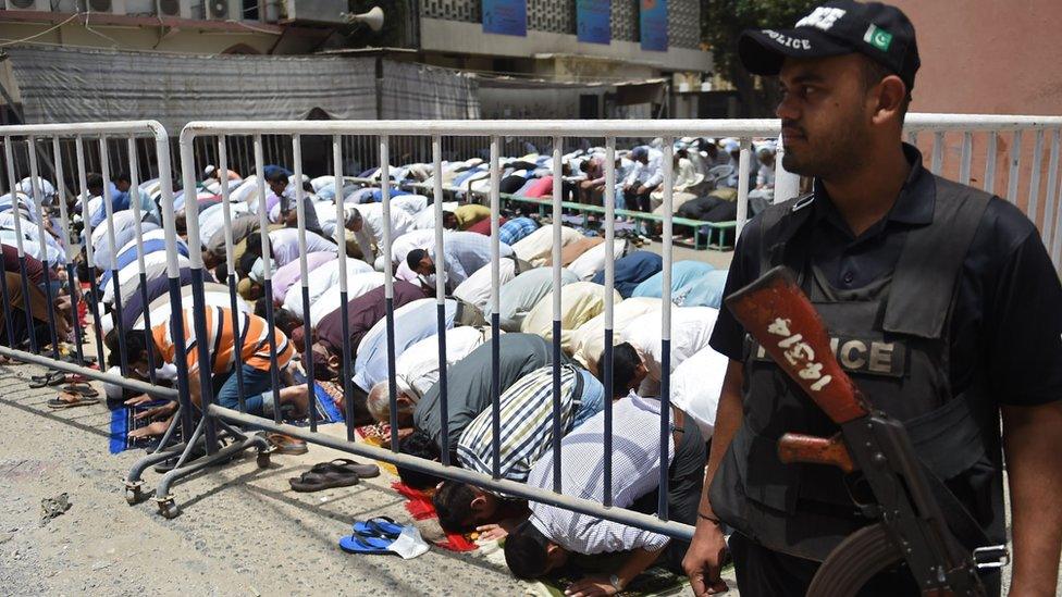 A Pakistani policeman armed with an AK-47 stands guard in front of a large group of praying men