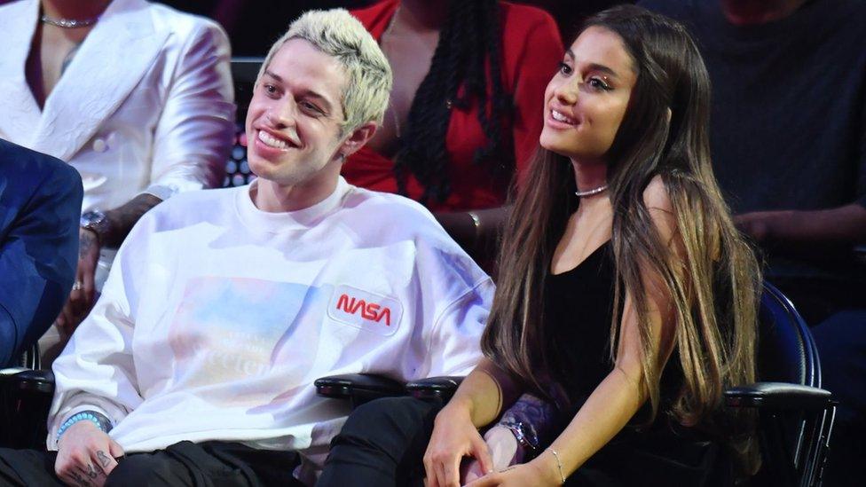 Pete Davidson and Ariana Grande look into the distance smiling.