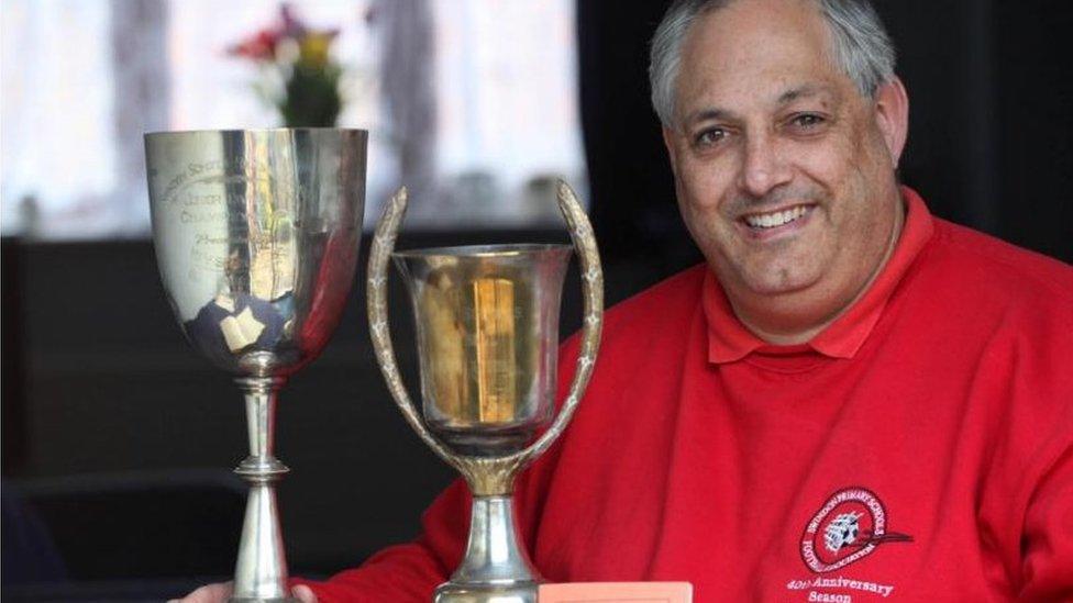 Man in red jumpers holds two football trophies