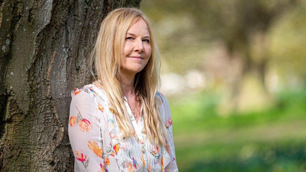 Sarah Class posing in front of a tree while wearing a white blouse with a colourful floral design