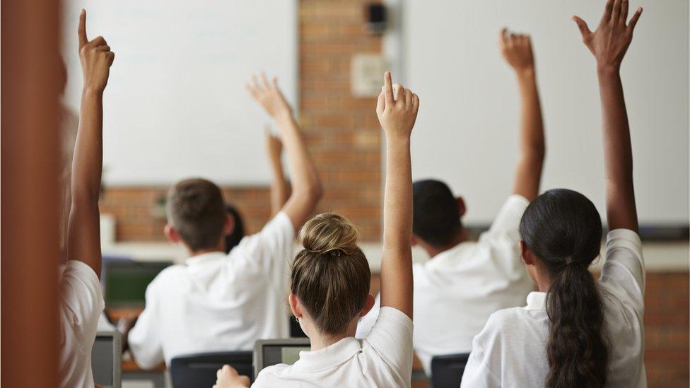 A group of pupils in a classroom seen from behind. All of the pupils have white shirts on, and all of them have their hands raised as if to answer a question