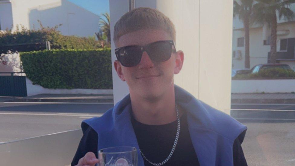 Jake Smith with sunglasses on, smiling at the camera wearing a black top and metal chain necklace