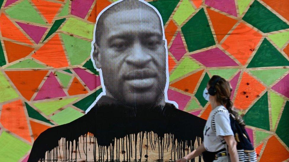 A woman walks past a boarded up store front with a George Floyd mural